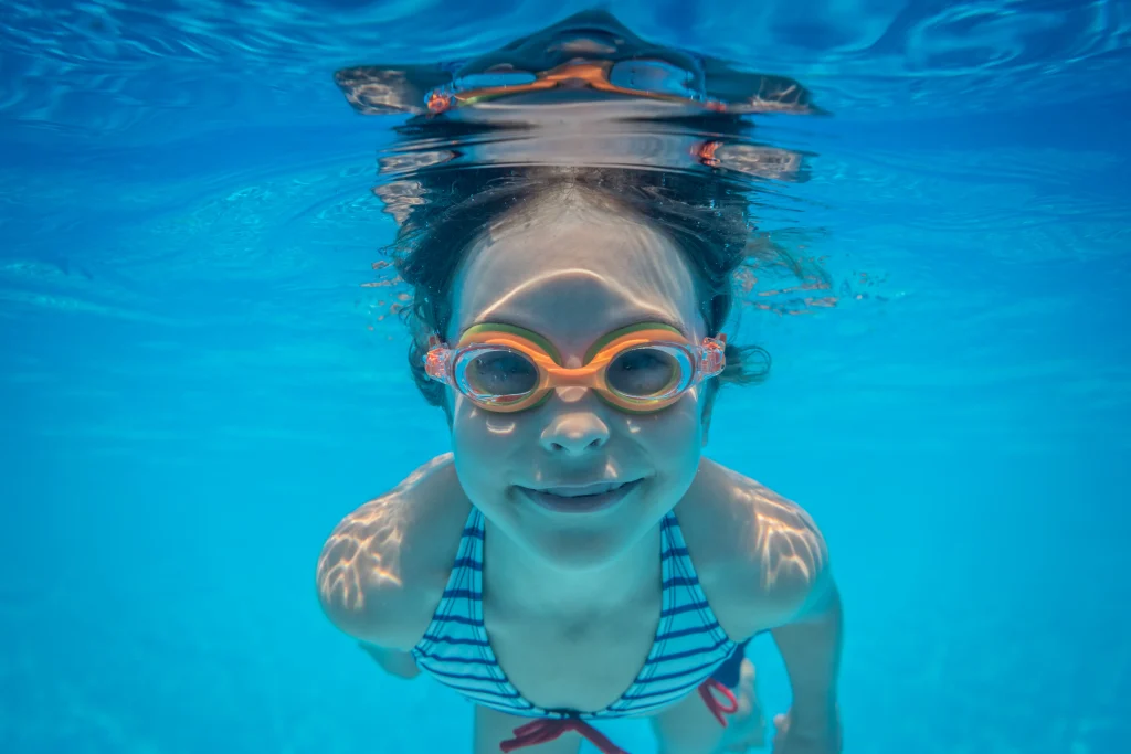 schwimmen lernen kinder: Kind taucht mit orangefarbener Schwimmbrille, ruhiges Lächeln, Wassergewöhnung