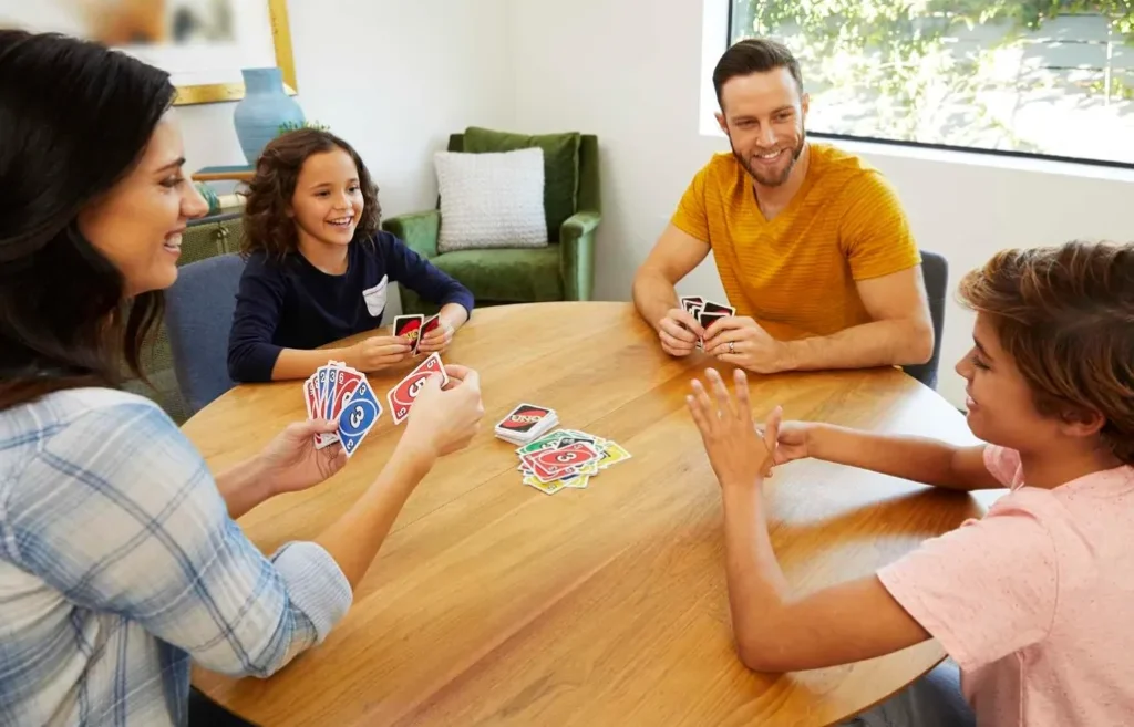 Familie spielt Uno am runden Holztisch; fröhliche Runde mit Kindern, bunte Uno-Karten liegen offen in der Tischmitte.