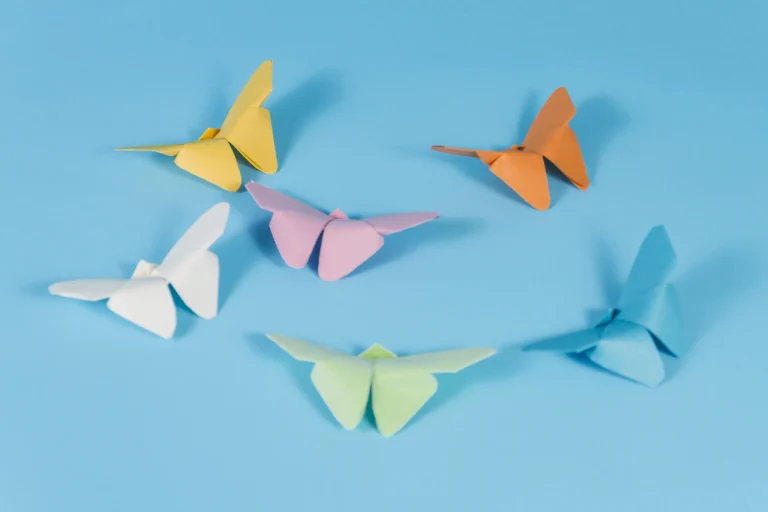 Sechs farbige Origami Schmetterlinge aus Papier auf blauem Hintergrund, in lockerer Kreisform angeordnet.