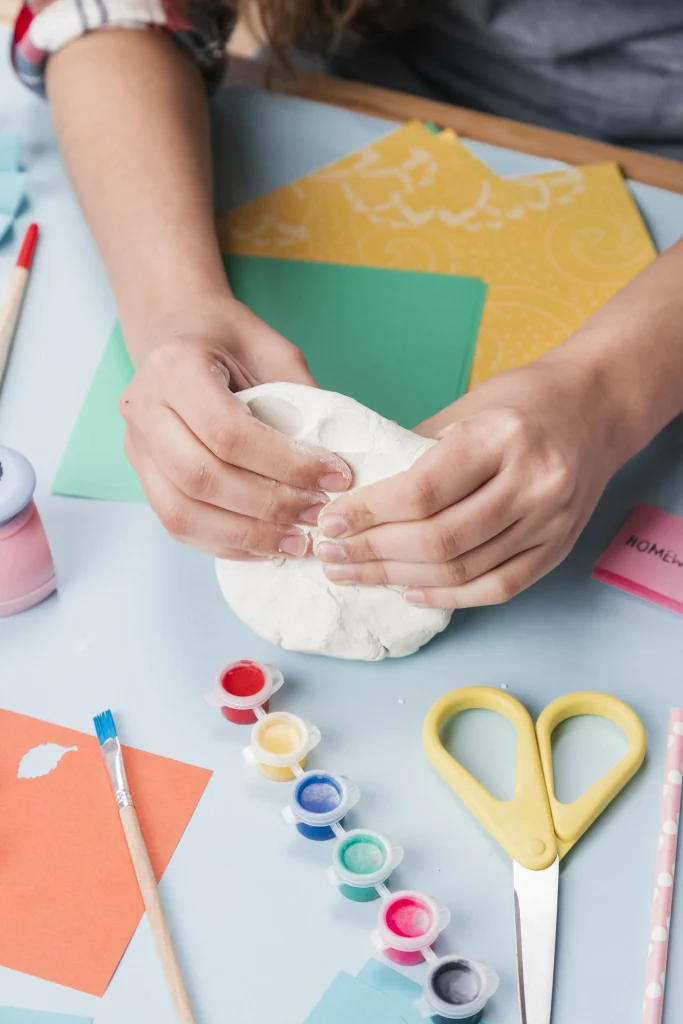 Kinderhände kneten weißen Salzteig; auf dem Tisch liegen Wasserfarben, Pinsel, Schere und Tonpapier.