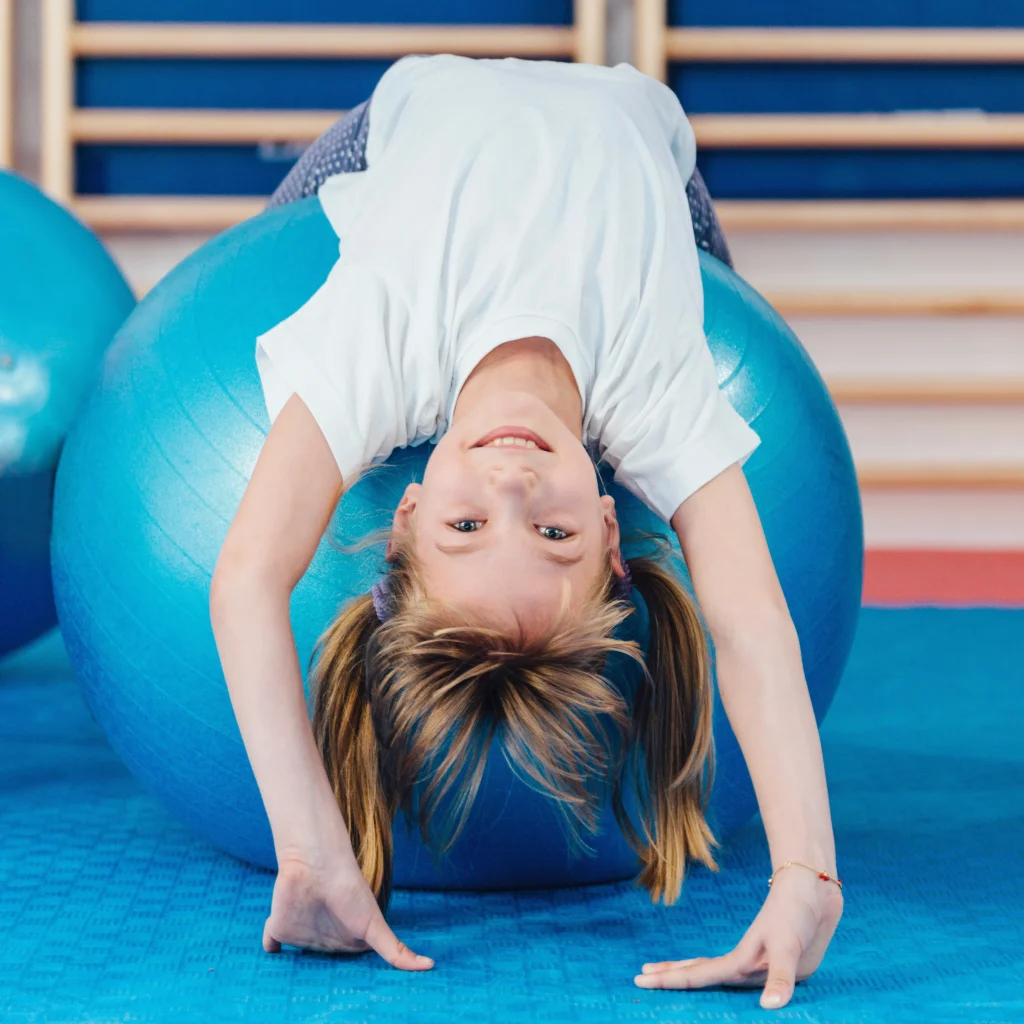 Kinderturnen in der Kita – Mädchen liegt rücklings über einem blauen Pezziball und stützt sich mit den Händen am Boden ab; Übung zur Rumpfstabilität und Balance