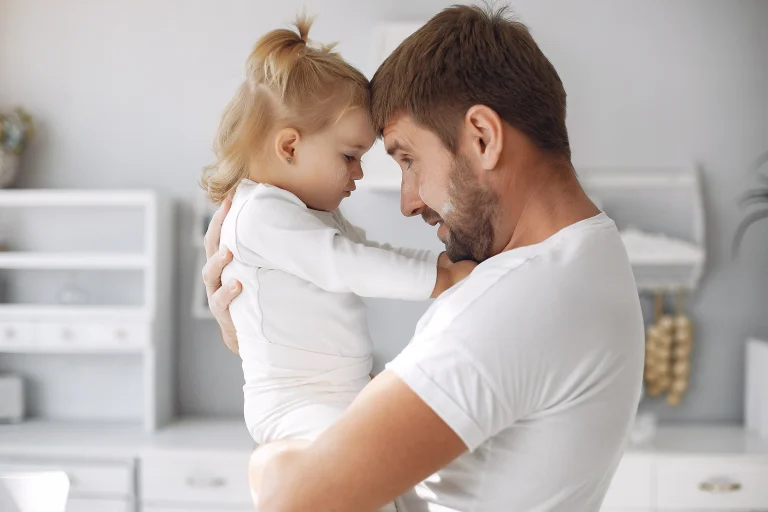 bedürfnisorientierte erziehung: Vater hält sein Kleinkind im Arm, beide berühren die Stirn aneinander in heller Küche – Nähe, Feinfühligkeit und Co-Regulation