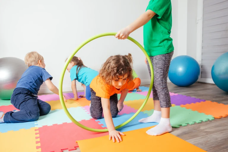 Kinderturnen in der Kita – Kind kriecht durch einen Hula-Hoop-Reifen auf bunten Turnmatten, angeleitet von anderen Kindern; Bewegungsparcours als Idee für Erzieher:innen