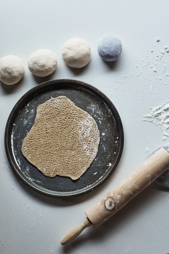 Ausgerollter Salzteig auf Backblech mit Nudelholz und Teigkugeln auf bemehlter Arbeitsfläche.