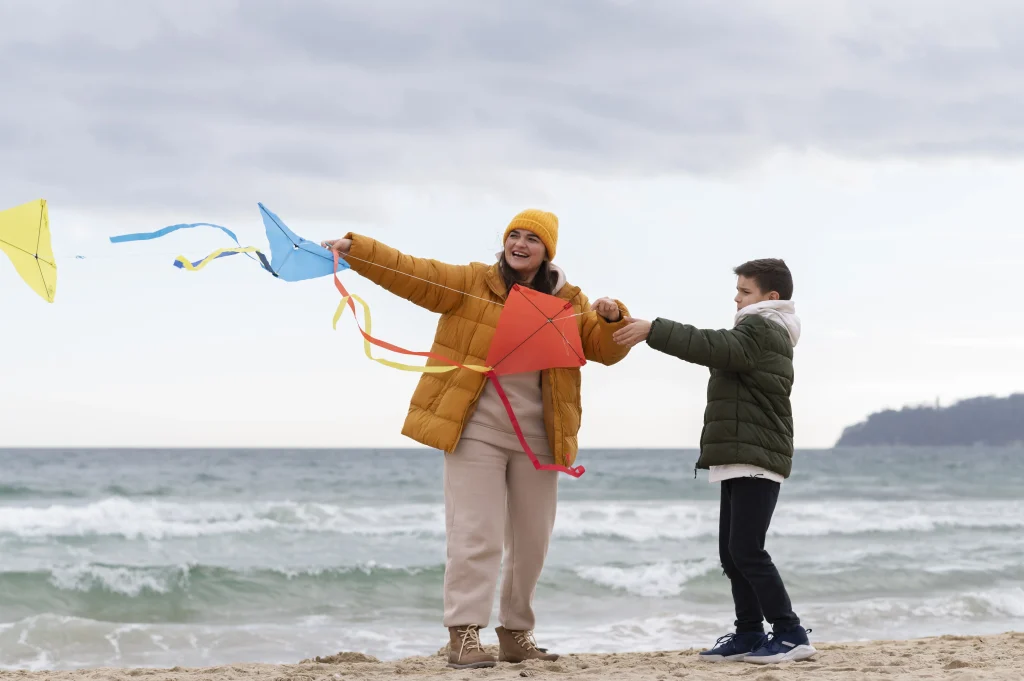 Mutter und Kind am Strand halten selbst gebastelte Rautendrachen mit buntem Schwanz im Wind – Drachen basteln und steigen lassen.
