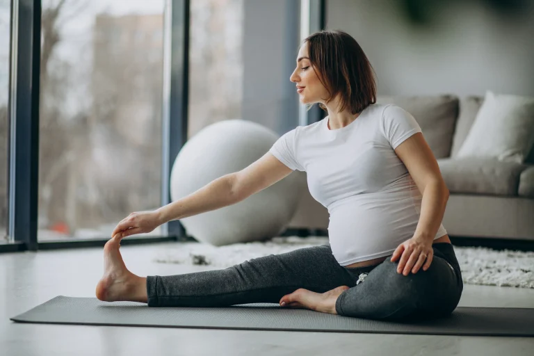 Schwangerschaftsyoga: Schwangere Frau sitzt auf der Matte in einer sanften seitlichen Dehnung (Janu-Sirsasana-Variation) in einem hellen Wohnzimmer.
