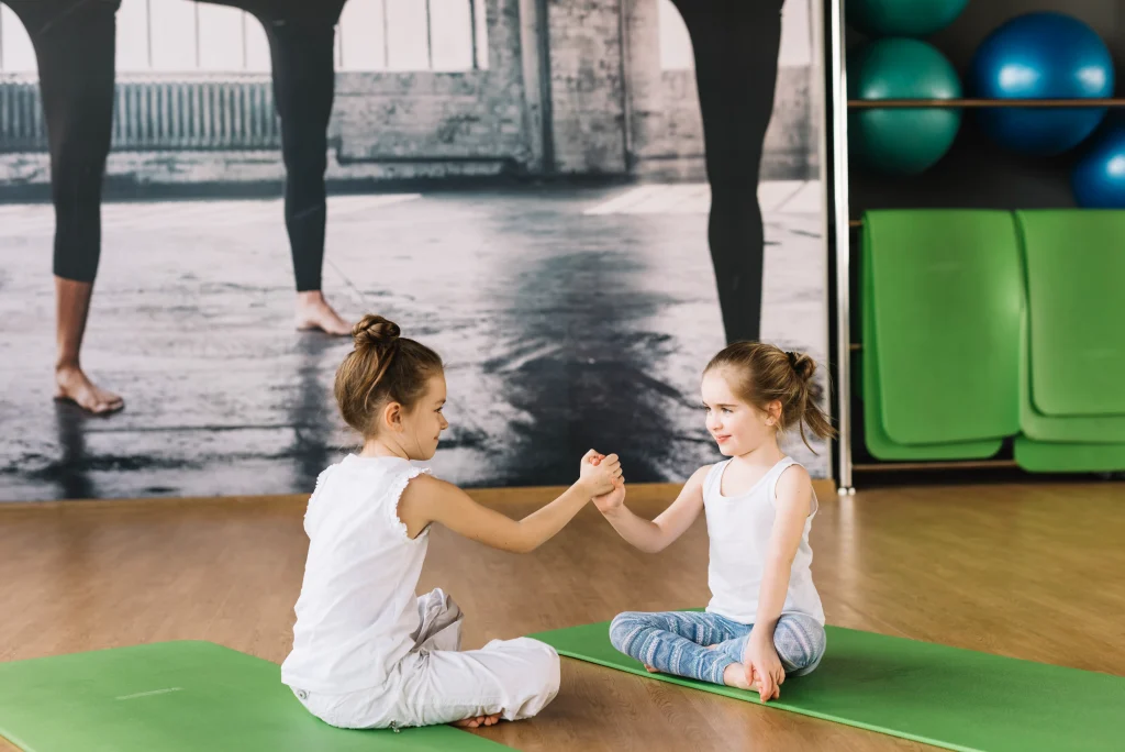 Kinderturnen in der Kita – zwei Kinder sitzen auf grünen Yogamatten und berühren sich zur Partnerübung an den Händen; Ritual für Fokus, Teamgefühl und Ruhe