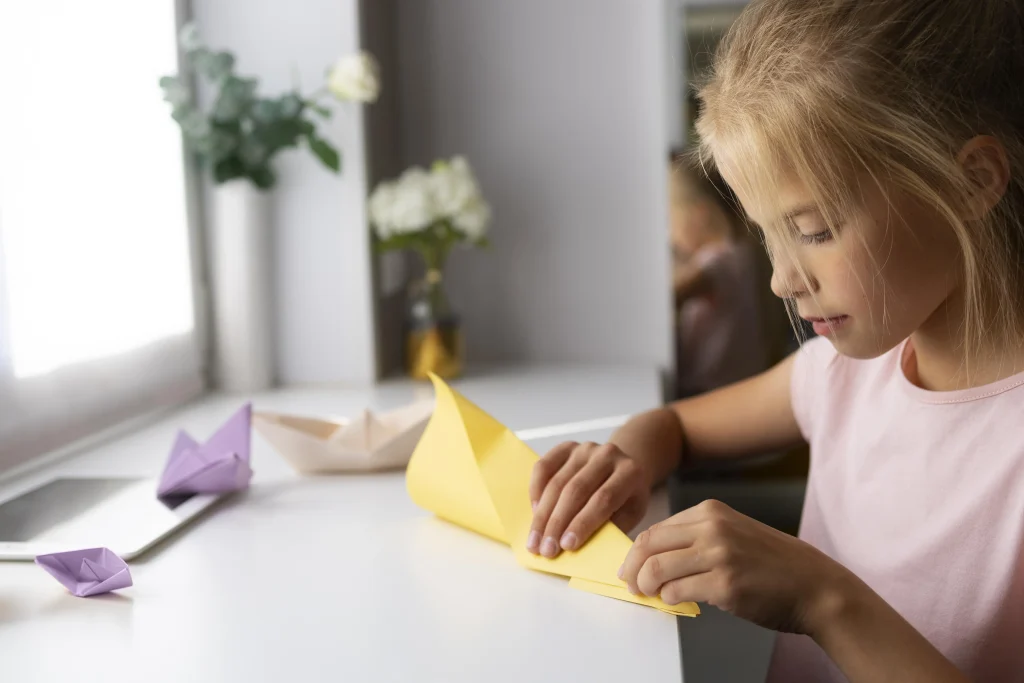 Kind faltet aus gelbem Papier einen Papierflieger am Schreibtisch – Papierflieger basteln in der Kita.