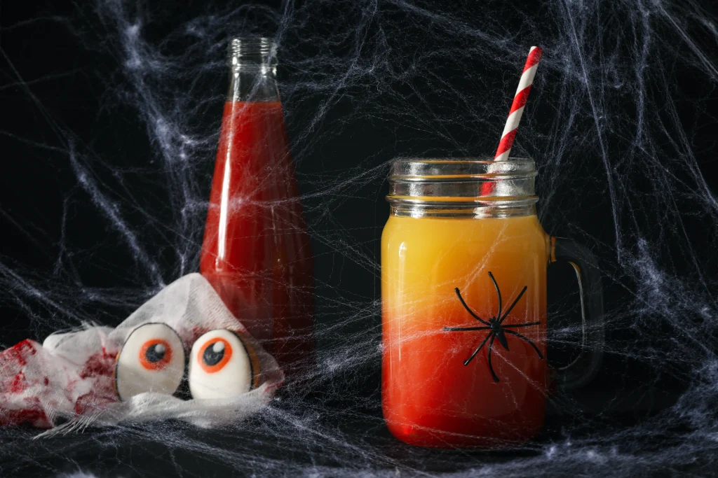 Halloween Deko basteln mit Kindern – gruseliger Kinderdrink im Glas mit Papier-Spinne, rote Limonade-Flasche und Zucker-Augen zwischen Spinnweben