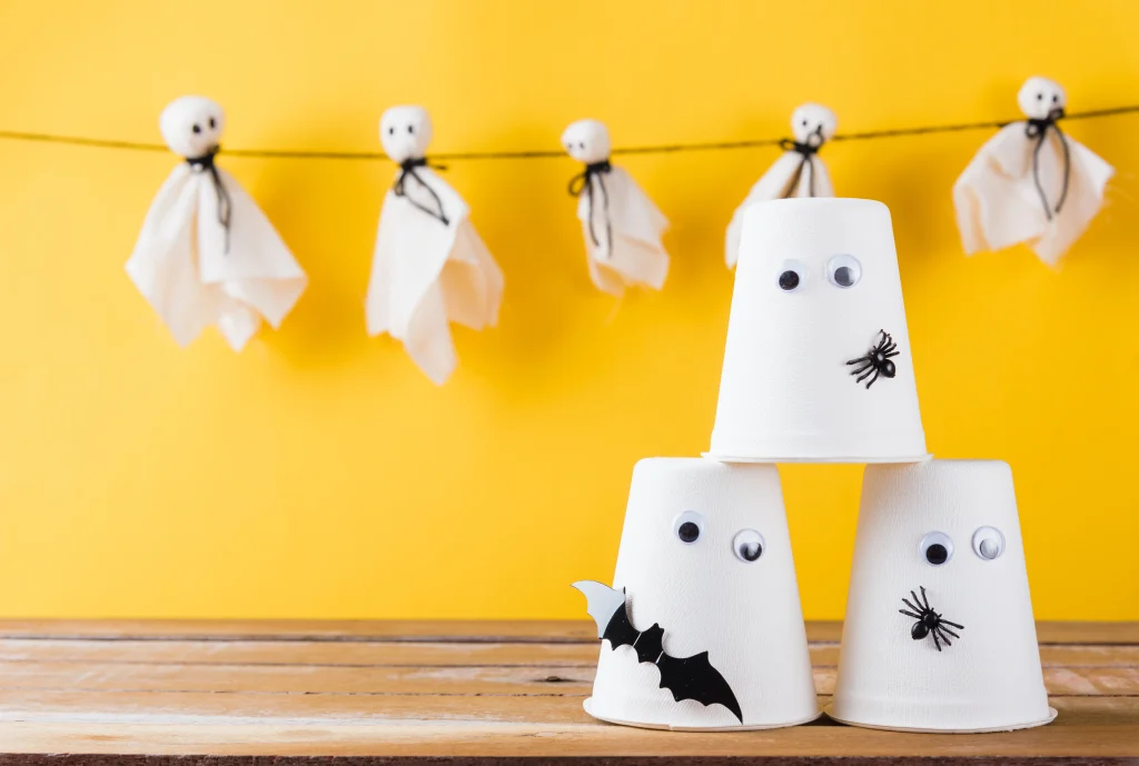 Halloween Deko basteln mit Kindern – Geistergirlande aus Stofftüchern und umgedrehte Becher mit Wackelaugen, Spinnen und Fledermaus vor gelbem Hintergrund