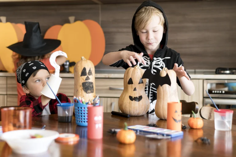 Halloween Deko basteln mit Kindern – zwei Kinder bemalen Kürbisse und gestalten Gesichter und Fledermaus-Deko am Tisch in der Küche