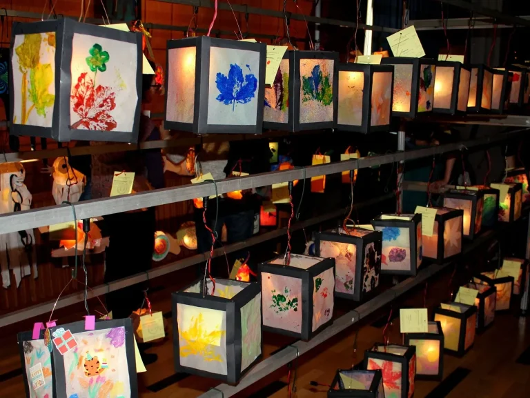 Reihen aus selbst gebastelten, quadratischen Papierlaternen mit bunten Blattdrucken hängen in einer Halle und leuchten warm mit LED-Lichtern – Laterne basteln in Kita und Grundschule.