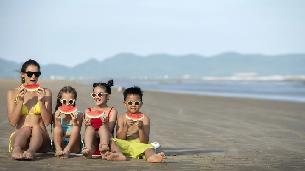 Erwachsene Begleitperson mit drei Kindern am Strand, alle essen Wassermelone – entspannte Familienzeit, ideal zur Planung von Kita-Schließtagen und Schulferien