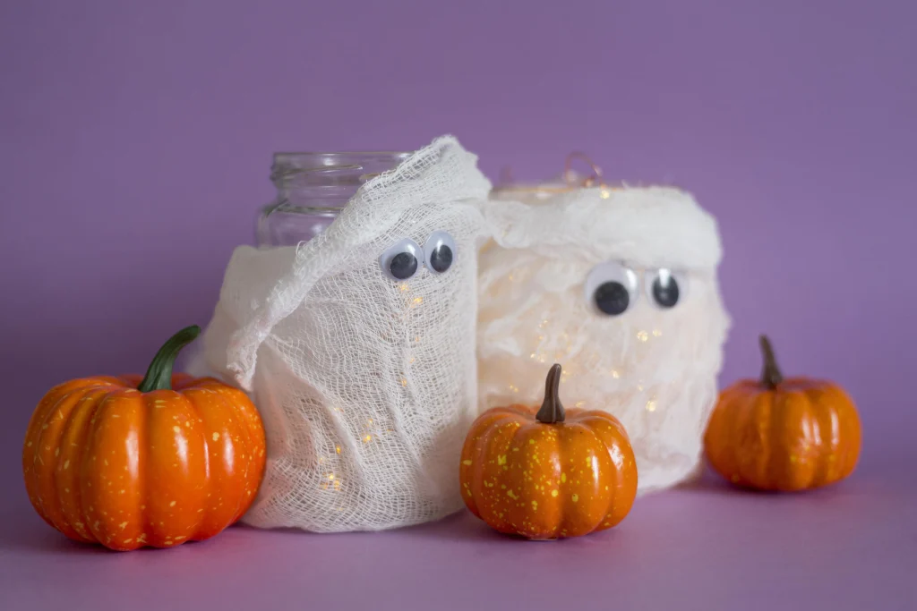 Halloween Deko basteln mit Kindern – Mumien-Windlichter aus Einmachgläsern mit Mullbinden, Wackelaugen und LED-Lichtern neben kleinen Zierkürbissen