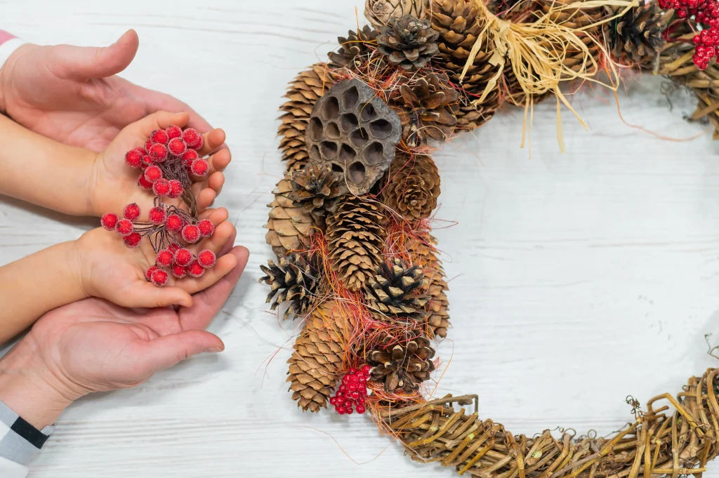 Kinder- und Erwachsenenhände halten rote Beeren als Dekoration für einen Adventskranz aus Zapfen und Naturmaterialien.