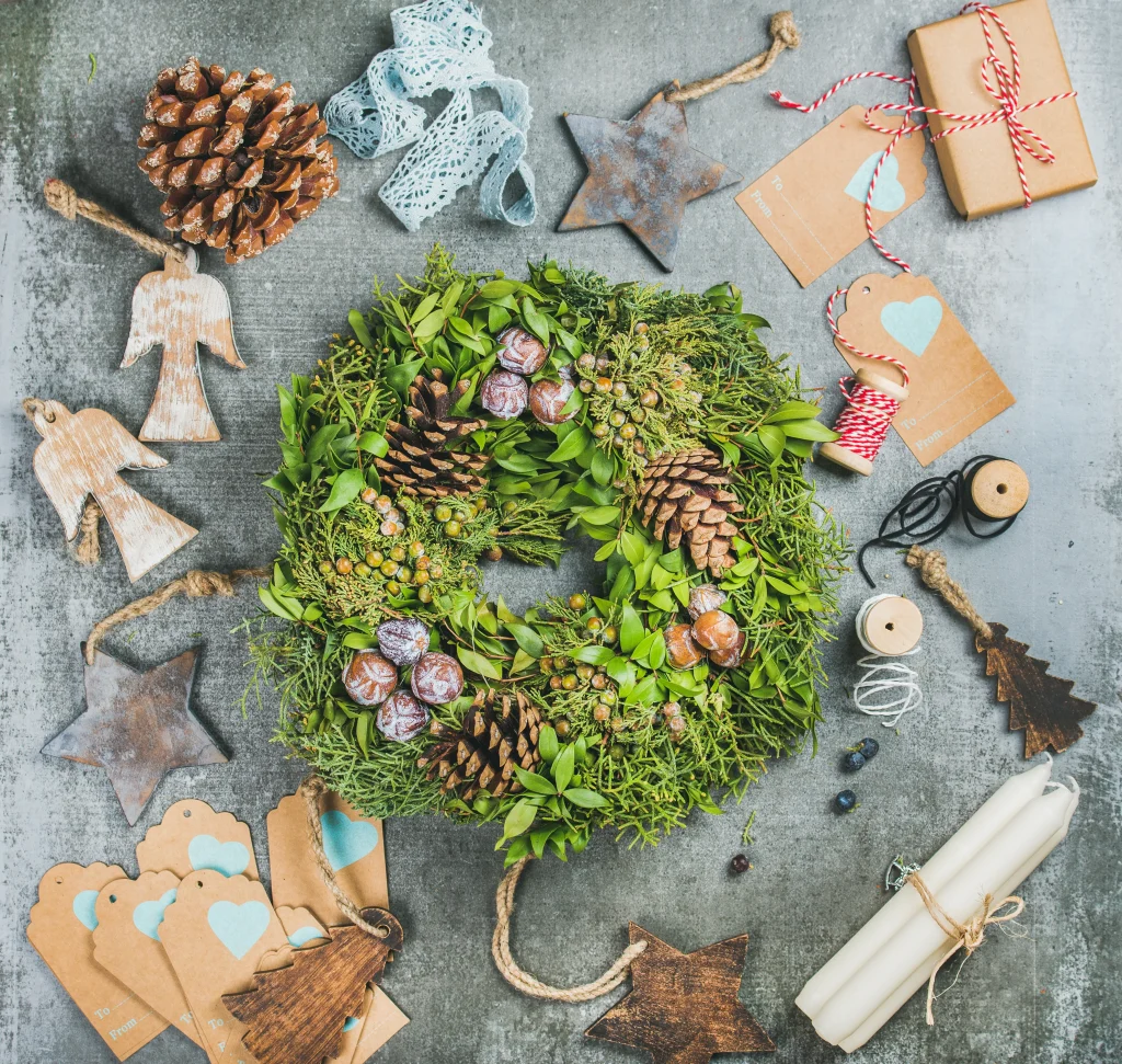 Grüner Adventskranz aus Naturmaterialien mit Zapfen und Beeren, umgeben von weihnachtlichen Bastelmaterialien wie Holzfiguren, Etiketten, Kerzen und Bändern.