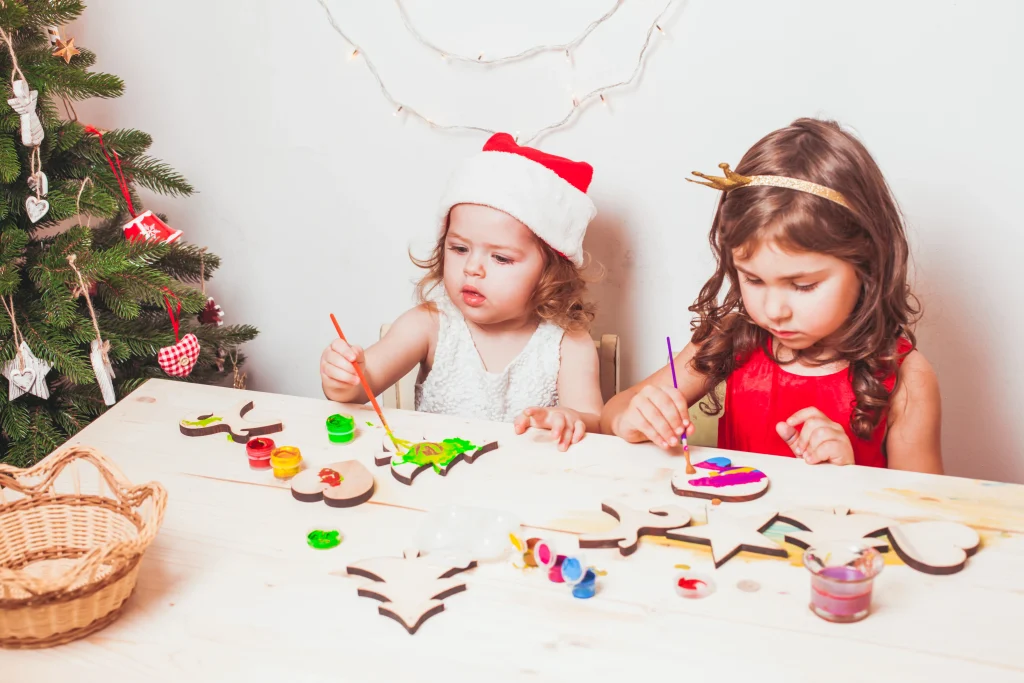 Kinder basteln Weihnachtsdeko mit Kindern in der Kita und bemalen weihnachtliche Holzanhänger am Tisch.