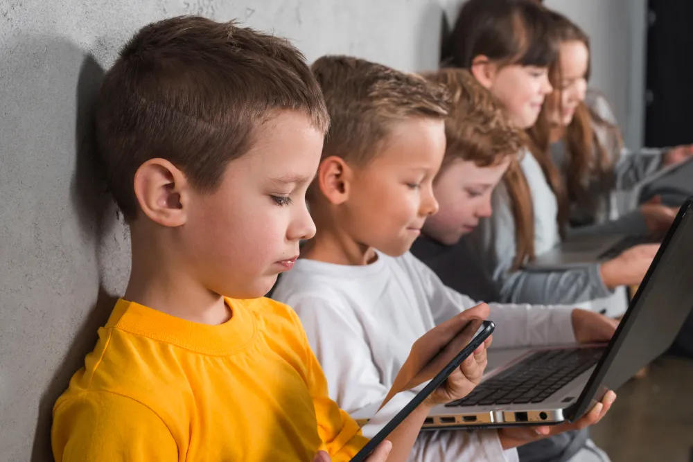 Kinder sitzen nebeneinander und nutzen Smartphones und Laptop – Medienkompetenz bei Kindern im digitalen Alltag