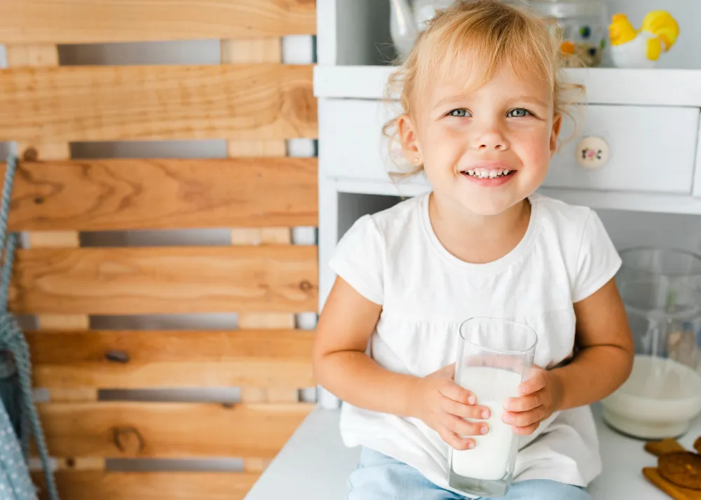 Kleinkind sitzt mit einem Glas Kuhmilch in der Küche und lächelt in die Kamera