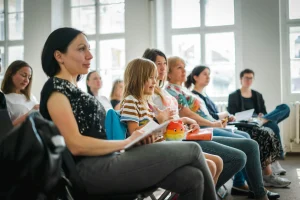 Erwachsene sitzen mit einem Kind bei einer gemeinsamen Veranstaltung in einem hellen Raum und halten Unterlagen in den Händen.